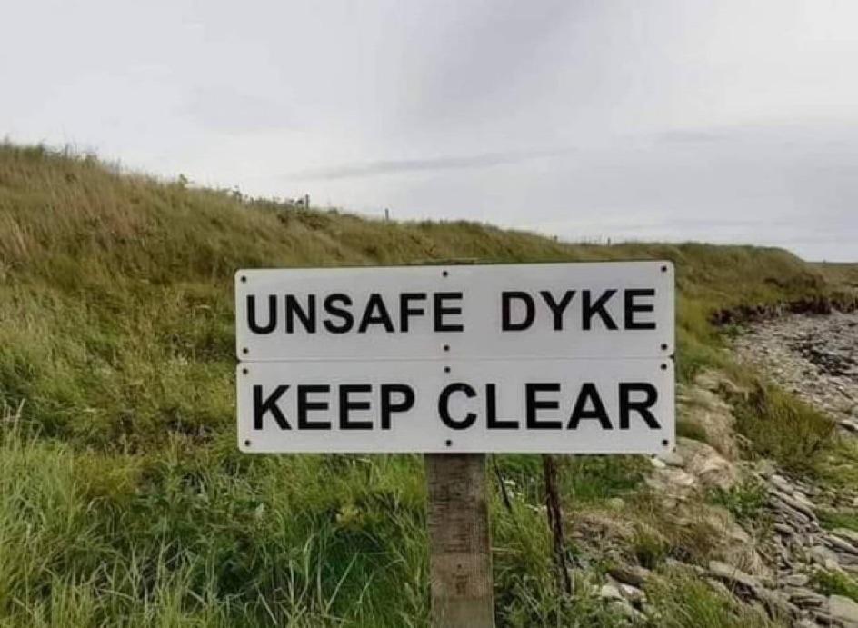 Photo of a sign on a grassy creek bank, reading "unsafe dyke keep clear"